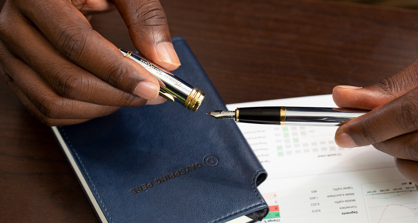 A man uncapping a fountain pen over a journal on a desk.